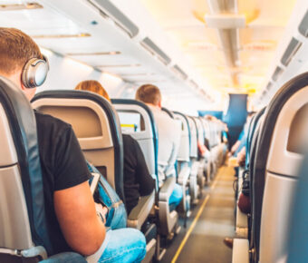 Economy class airplane cabin with passengers seated in armchair-style seats along a central aisle.