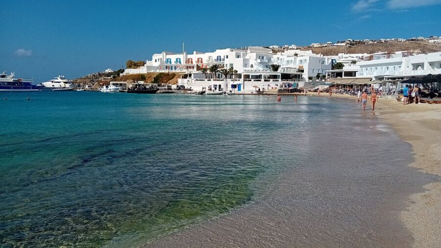 Platis Gialos Beach in Mykonos, Greece, showcasing white buildings and visitors relaxing on the sandy beach.
