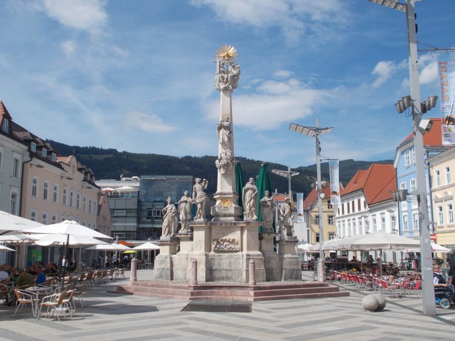 Gilded sunburst-topped monument and column with statues in historic plaza with outdoor cafes.