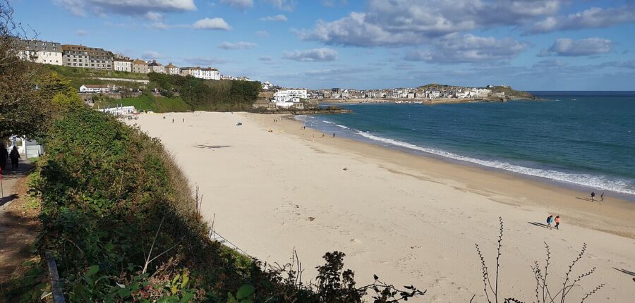 Porthminster Beach in Cornwall, England, with people walking along the shore and a hill in the background.
