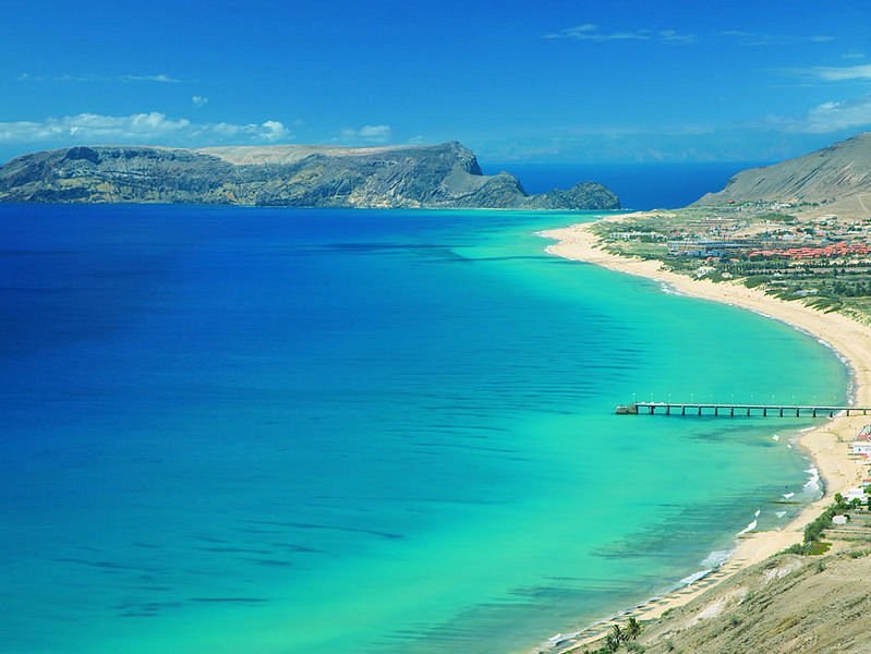 Porto Santo beach with clear blue water and majestic mountains rising in the background.
