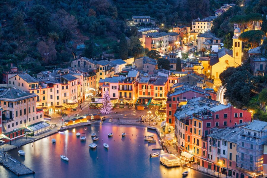 Portofino, Italy harbor at dusk with illuminated waterfront, boats, and pastel village buildings.