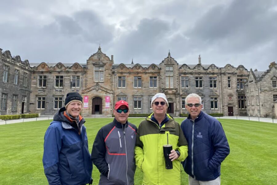 Four men in jackets and hats stand smiling on a grassy lawn with a grand historic stone building behind them on a Private St. Andrews Town Walking Tour