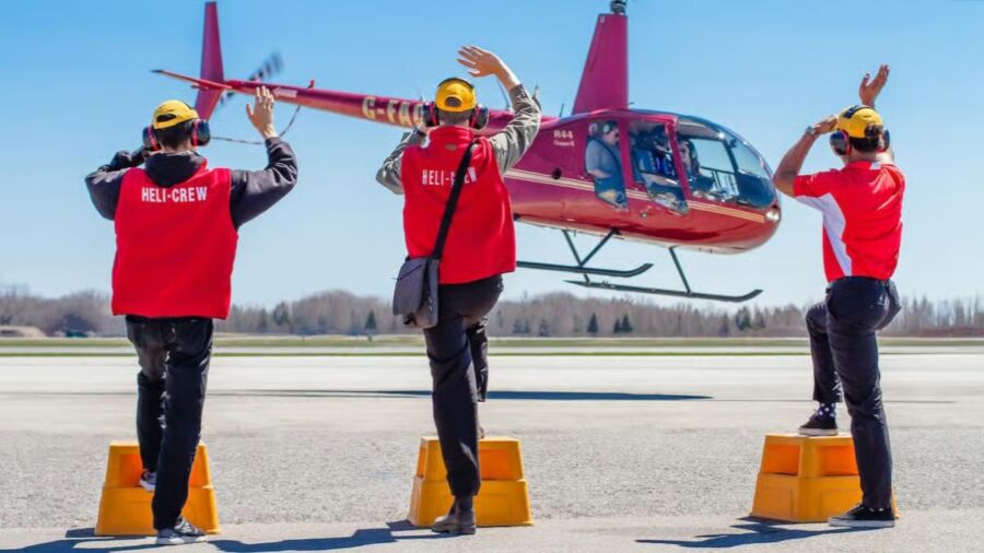 Three crew members in red vests are directing a red helicopter on a sunny tarmac. They stand on yellow platforms, gesturing with raised arms.
