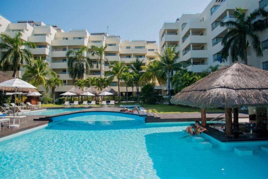 The pool area at Privilege Aluxes hotel features lounge chairs and umbrellas, set against a backdrop of Isla Mujeres.
