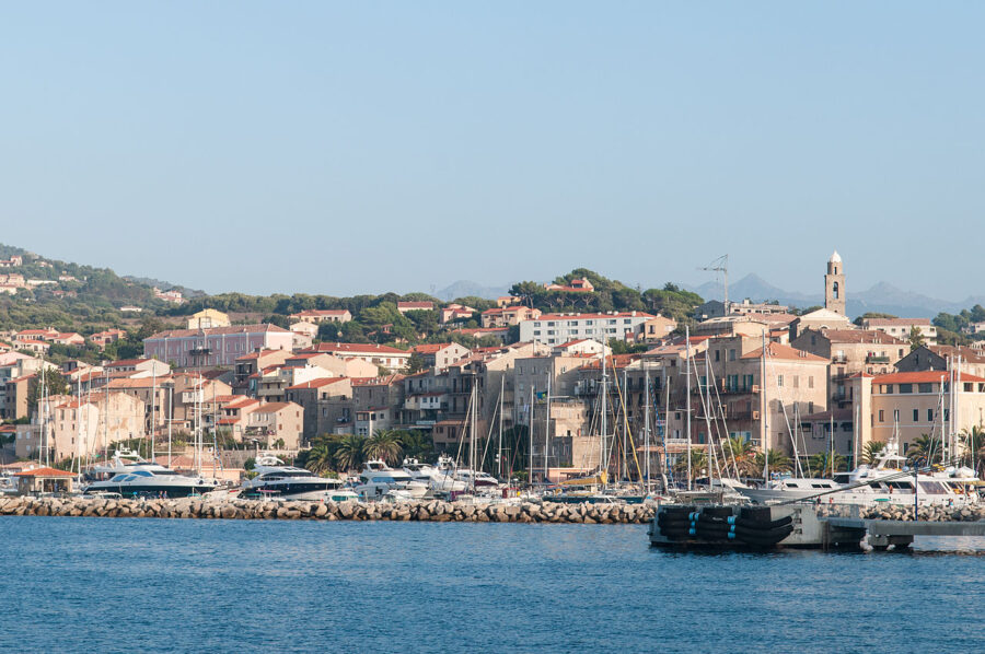 Tranquil waters of Propriano, Corsica, France, surrounded by lush greenery and distant hills under a bright sky.