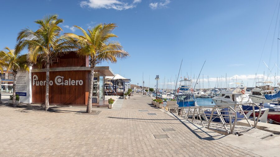 Puerto Calero marina promenade with yachts, palm trees, and modern harbor buildings on a sunny day.