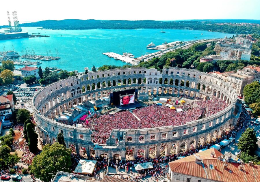 Aerial view of Pula Arena crowd celebrating, with Adriatic Sea in background.