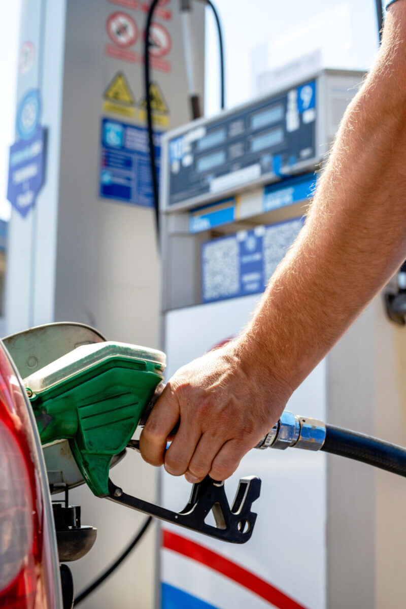 Close-up of a hand holding a green gas pump nozzle, refueling a car at a station. The background shows a fuel dispenser with various signs.