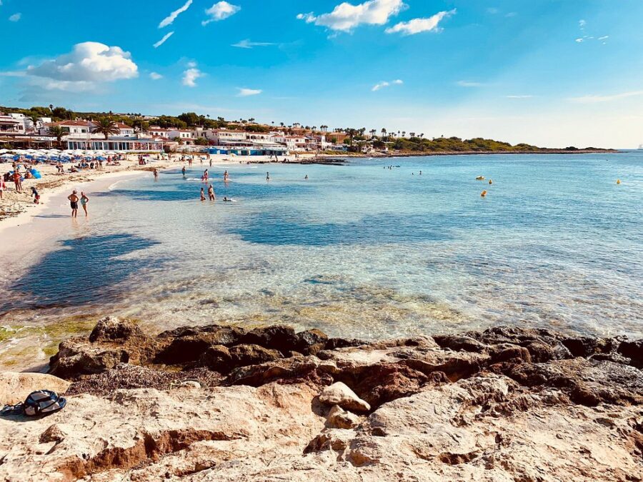 Bustling beach scene at Punta Prima, Menorca, with numerous people relaxing and playing in the sand and water.