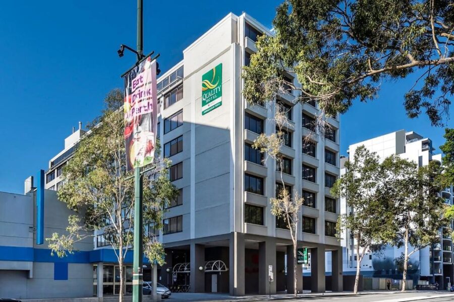 Quality Hotel in downtown Perth city street with modern facade, signage, trees, and clear sky.