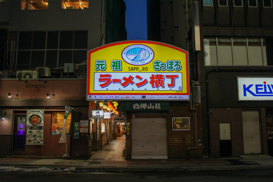 Nighttime Sapporo ramen alley entrance with glowing Japanese sign, lanterns, and wet winter street.
