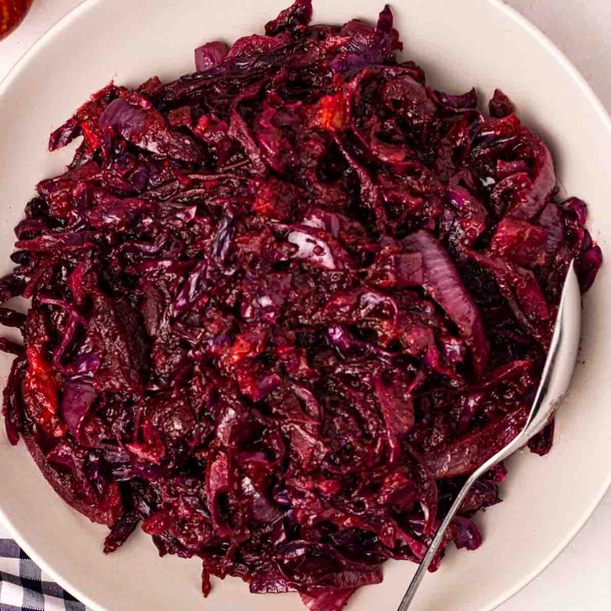Vibrant shredded braised red cabbage in white bowl with serving spoon, inviting and colorful.