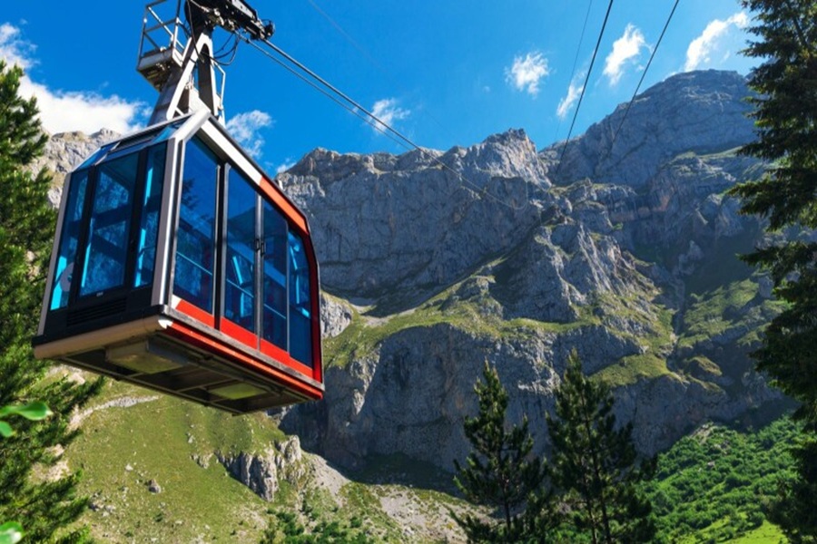 Red gondola cable car over alpine cliffs and conifer forests under a clear blue sky.