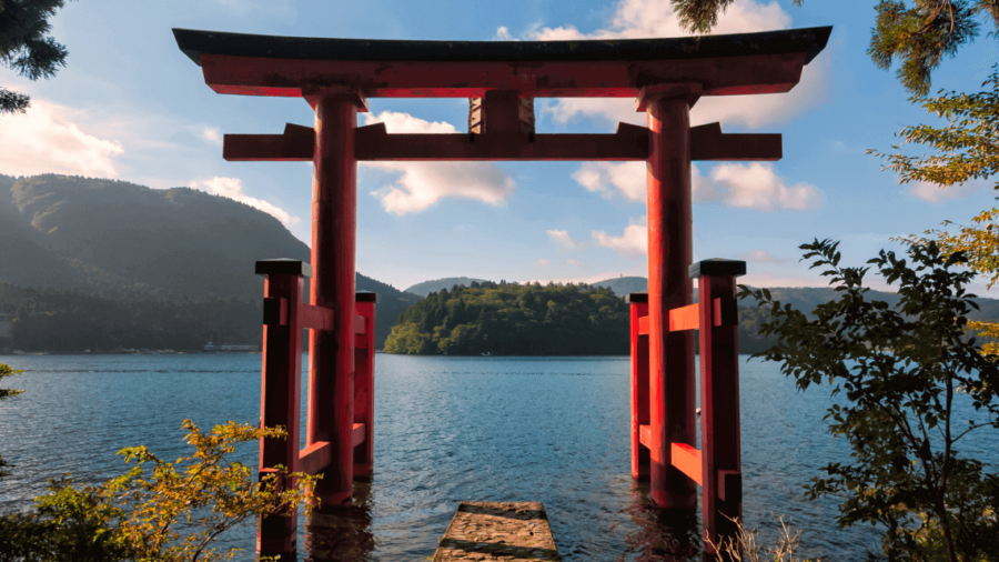 Red torii gate on tranquil Japanese lake with mountains, forest island, and blue sky.