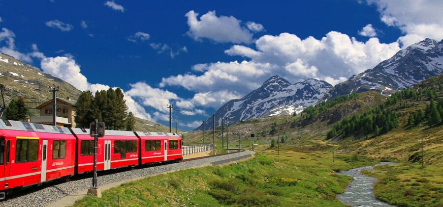 Red passenger train winding through alpine valley with mountains and blue sky.