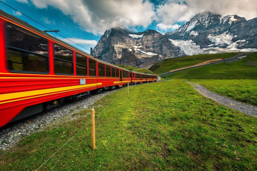 Red and yellow train in a stunning mountainous landscape with snow-capped peaks and lush greenery.