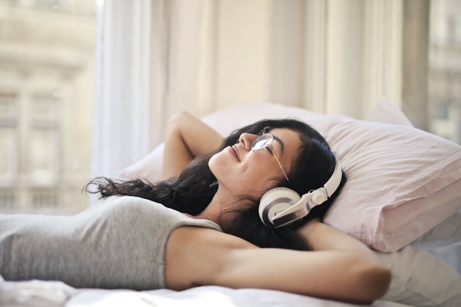 Young woman relaxing on bed with headphones, enjoying music in a bright, serene room.