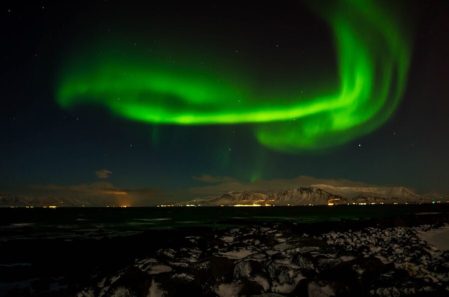 Vibrant green aurora borealis dances across a dark night sky over a snow-covered landscape of Reykjavik and distant mountains, creating a serene and magical atmosphere.