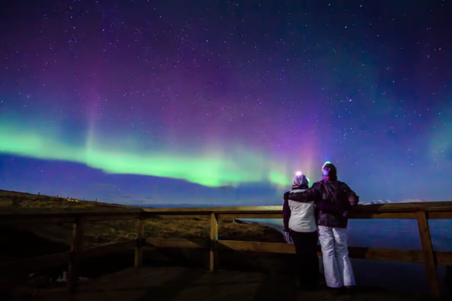 Two people on a wooden deck gazing at the vibrant aurora borealis during the Reykjavik Premium Northern Lights Tour