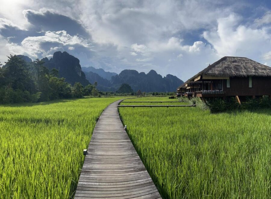 Scenic wooden boardwalk through lush rice fields toward Vang Vieng mountain bungalows in Laos.