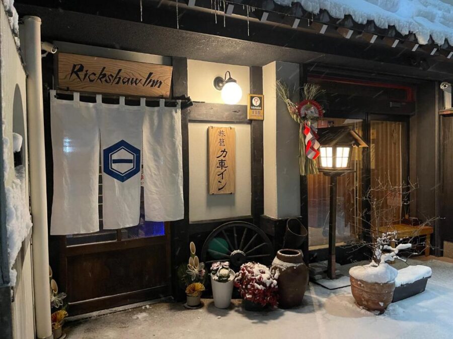 Snowy Japanese ryokan entrance at night with warm lanterns, noren curtain, and winter decorations.