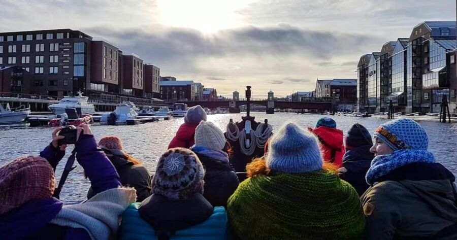 Group of bundled-up people on a boat admire a sunny, wintery harbor scene with modern buildings and yachts, conveying a sense of exploration and camaraderie on a River Nidelva Private Boat Guided Sightseeing Tour