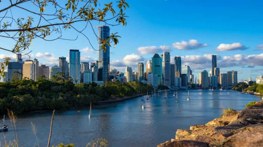 Brisbane riverfront city skyline with modern skyscrapers, boats, and trees at sunset.