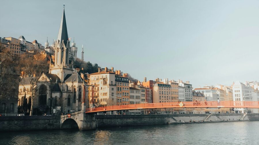 Lyon riverside with red pedestrian bridge, Gothic church spire, and reflective Rhone River.
