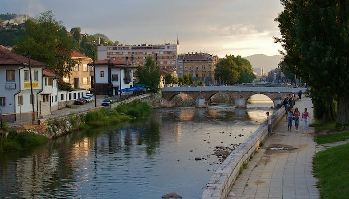 Peaceful Miljacka River promenade with stone bridge, historic buildings, minaret and evening walkers.