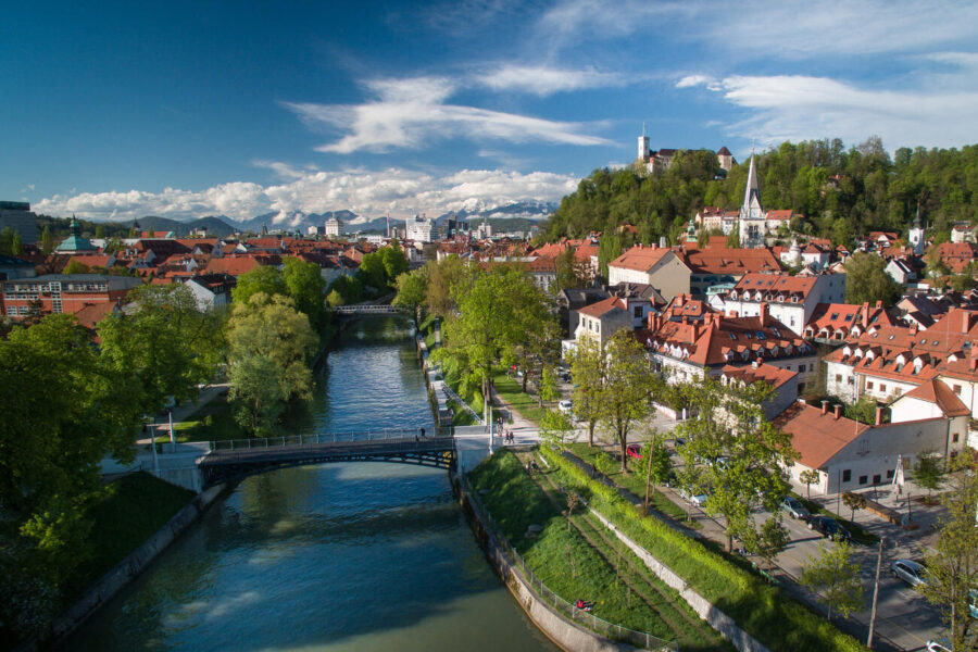 Sunlit European riverside town with arched bridge, red-tiled rooftops, church spire on wooded hill.