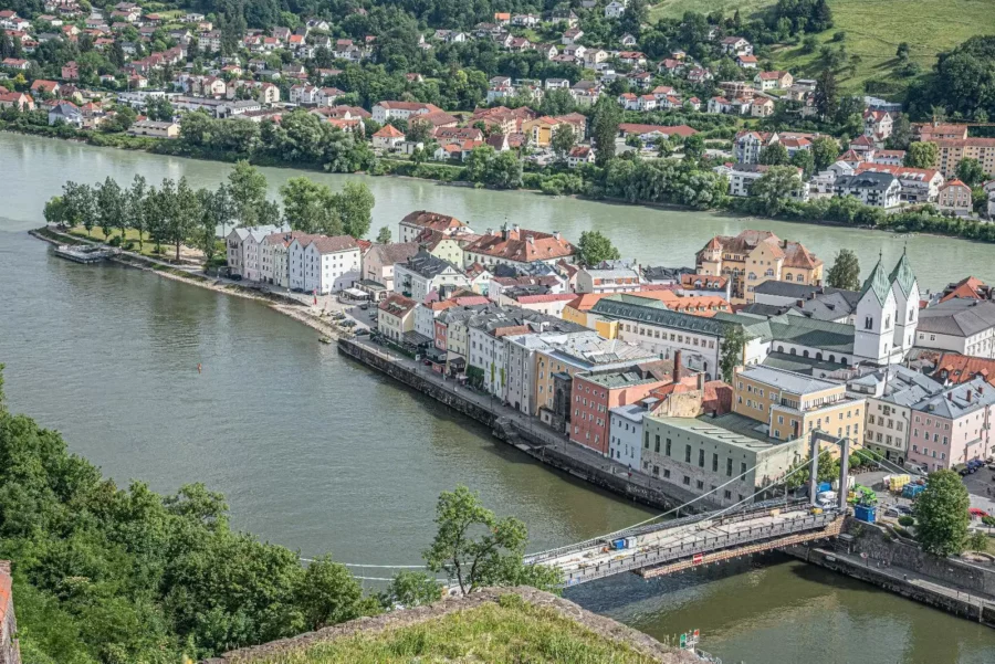 Scenic European riverside town with historic buildings, suspension bridge, and green hills in background.