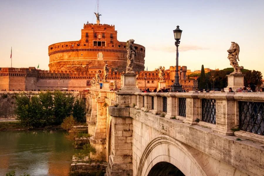 Historic stone fortress and angel statues on riverside bridge at sunset.