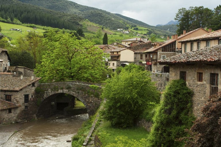 Medieval riverside village with stone arch bridge, terracotta roofs, and lush green hills.