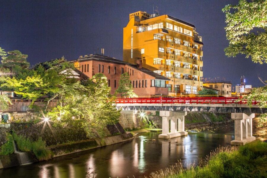 Night river in Japanese city with illuminated bridge, modern hotel, reflections, and lush green trees.