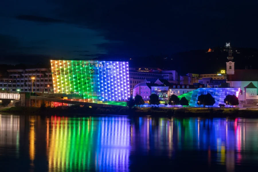 Colorful LED-lit modern riverside buildings and pavilion reflecting on river in nighttime cityscape.