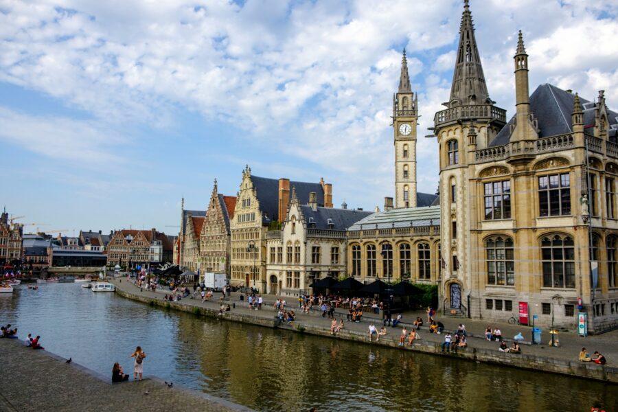 Ghent canal and quay with historic buildings, clock tower, boats, and people on the waterfront.