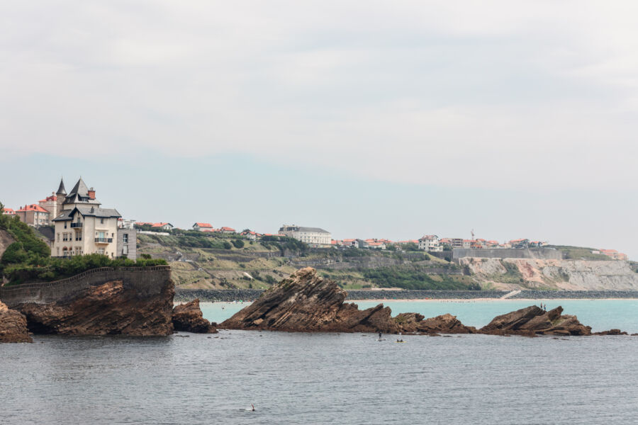 Rocher de la Vierge, French Basque Country  coastal landscape with a grand house set on a rocky cliff, overlooking calm sea waters. Jagged rocks protrude from the water under a cloudy sky.
