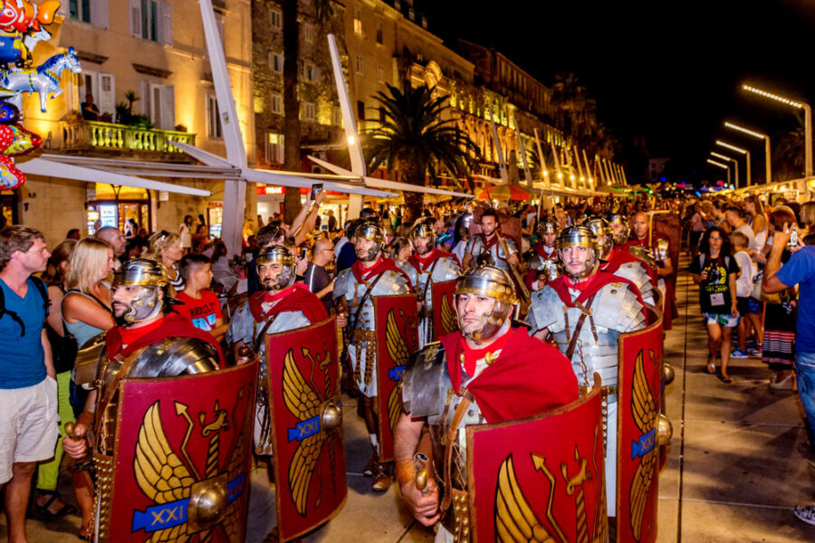 Roman soldiers parade at a vibrant, festive Mediterranean night with engaged crowd.