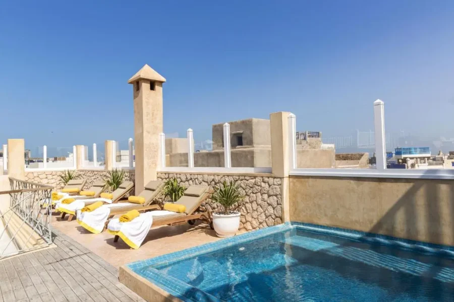 Rooftop plunge pool terrace with sun loungers, plants, and city skyline views on a sunny day.