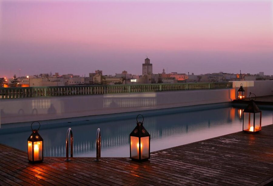 Luxury rooftop pool at dusk with lanterns overlooking historic city skyline and sunset sky.