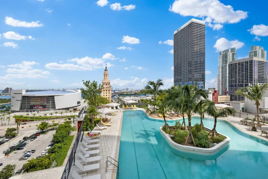 Sunny rooftop pool with palm trees overlooking city skyline and waterfront high-rises.