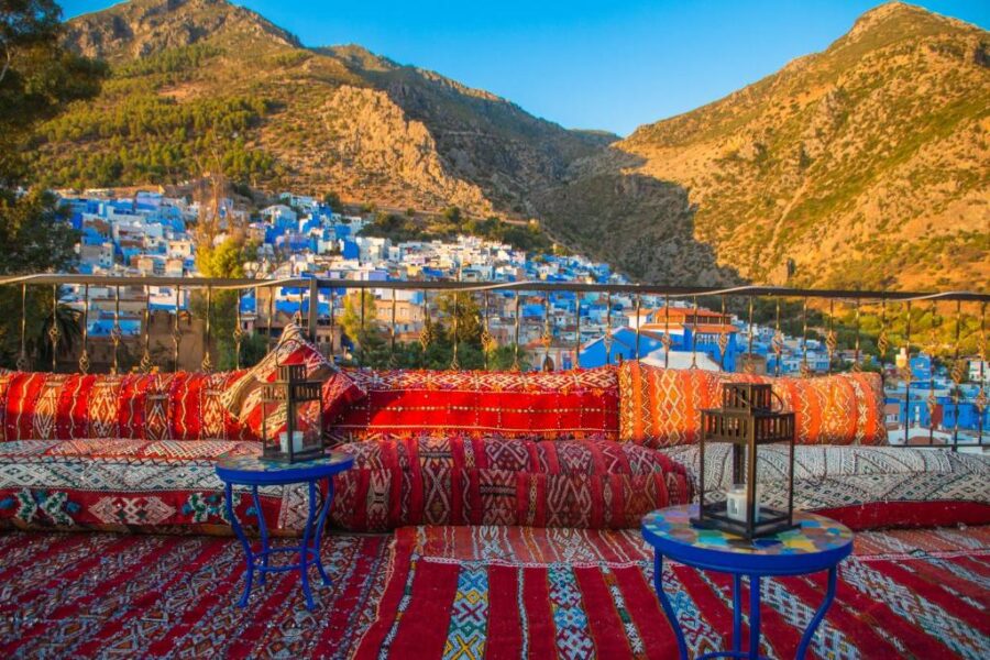 Colorful Moroccan rooftop terrace overlooking blue hillside town and surrounding rugged mountains at sunset.