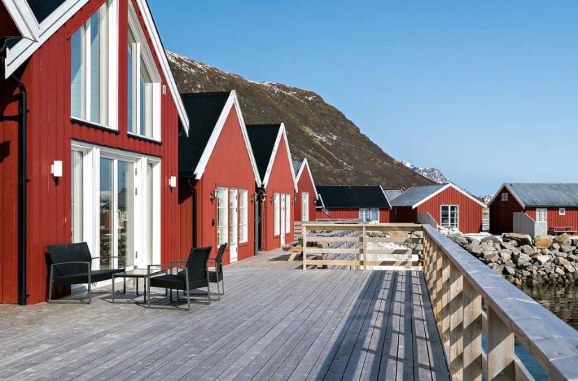 Rows of red wooden cabins in Rorbu i Lofoten with large windows line a waterfront deck. Black chairs and a table sit under a clear blue sky, with distant mountains.