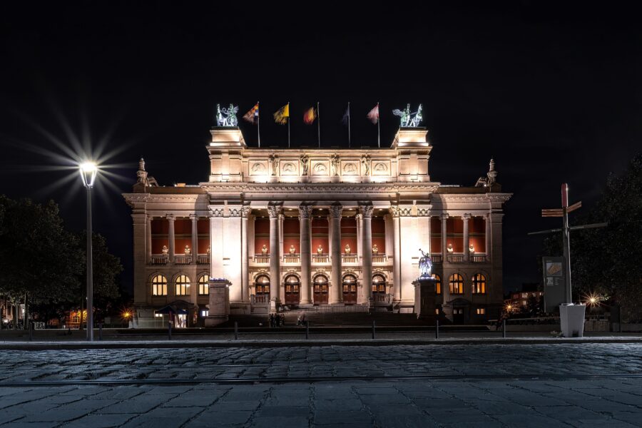 Night view of the Royal Museum of Fine Arts Antwerp, brightly lit and featuring various country flags in front.
