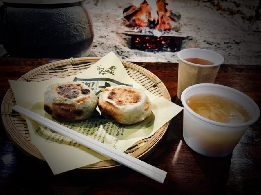 Pan-fried dumplings on bamboo platter by fire, with chopsticks and warm drinks.