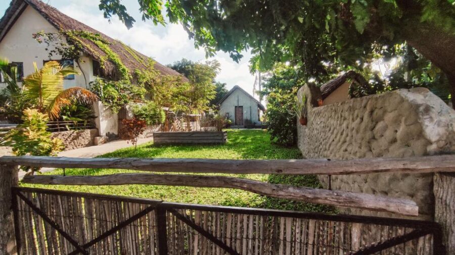Rustic thatched beach cottage with wooden gate, tropical garden, stone wall and sunlit green lawn.