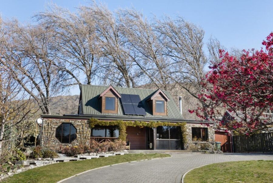 Rustic stone cottage with green roof, solar panels, garden landscaping, and pink flowering tree.