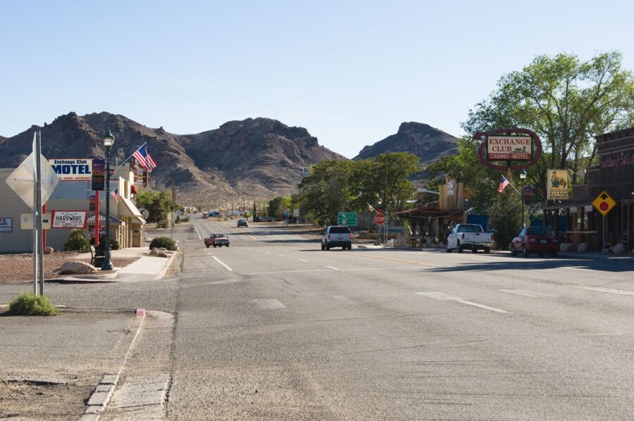 Small rustic town streetscape with mountains, Exchange Club Motel, shops, vehicles, and flags.