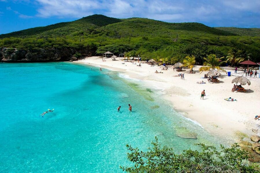 Clear waters and sunbathers populate the beach at Sabana Westpunt, Curacao, under a bright blue sky.
